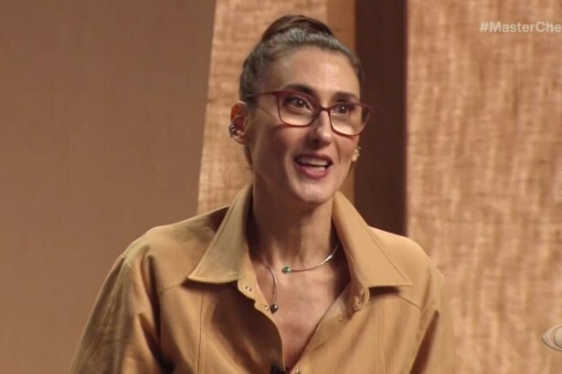 A chef e jurada Paola Carosella em um close-up, sorrindo e falando em uma cena do programa "MasterChef Brasil". Ela usa óculos de armação vermelha, uma camisa bege e um colar. O fundo do estúdio é de painéis de madeira. O logo da Band e a hashtag #MasterChefBR estão visíveis na tela.