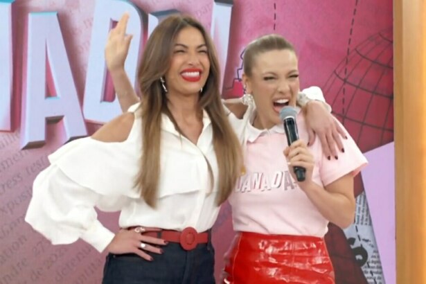 A imagem mostra as apresentadoras Patrícia Poeta e Beatriz Reis no palco do programa "Encontro". Patrícia Poeta, à esquerda, usa uma camisa social branca de mangas longas com ombros à mostra, calça jeans e um cinto vermelho, e está com a mão na cintura, acenando. Beatriz Reis, à direita, veste uma camiseta rosa e uma saia de couro vermelha, e segura um microfone com a mão direita, com os braços ao redor do pescoço de Patrícia. As duas estão sorrindo, com a boca aberta.
