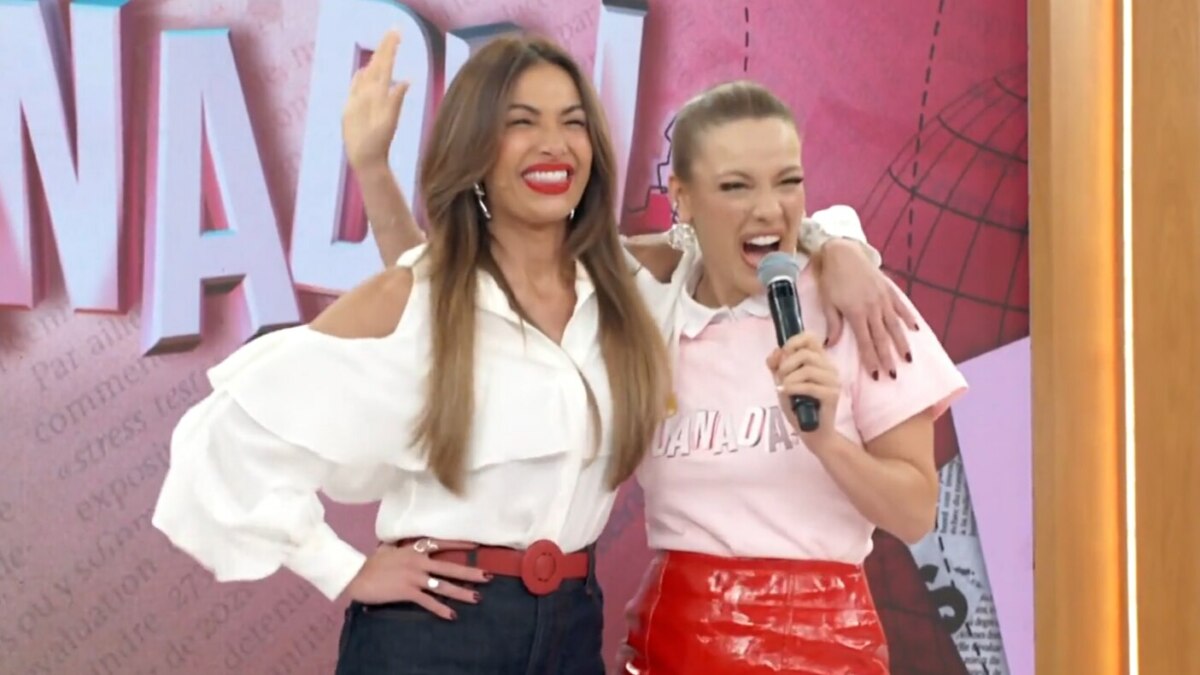 A imagem mostra as apresentadoras Patrícia Poeta e Beatriz Reis no palco do programa "Encontro". Patrícia Poeta, à esquerda, usa uma camisa social branca de mangas longas com ombros à mostra, calça jeans e um cinto vermelho, e está com a mão na cintura, acenando. Beatriz Reis, à direita, veste uma camiseta rosa e uma saia de couro vermelha, e segura um microfone com a mão direita, com os braços ao redor do pescoço de Patrícia. As duas estão sorrindo, com a boca aberta.