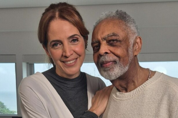 A jornalista Poliana Abritta e o cantor Gilberto Gil posam juntos em um ambiente claro. Poliana tem o braço sobre o ombro de Gilberto, e ambos sorriem levemente para a câmera. Ao fundo, grandes janelas revelam uma vista para o mar. A imagem é de uma entrevista para o programa "Fantástico".