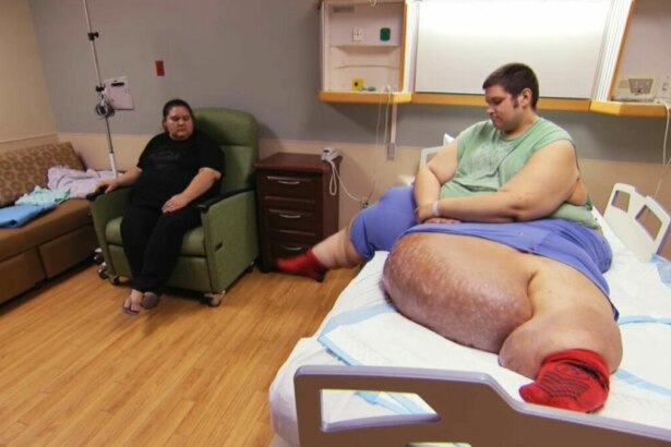 Um homem deitado em uma cama de hospital, com uma perna extremamente inchada, usa uma camisa verde e meias vermelhas. Uma mulher está sentada em uma poltrona próxima. O nome do arquivo o identifica como J.T., um paciente do programa 'Quilos Mortais'.