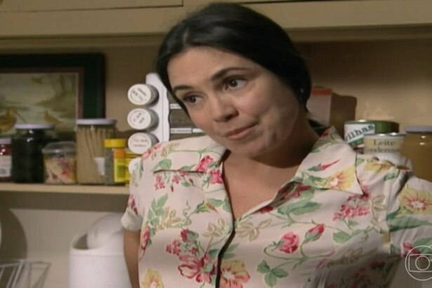 A atriz Regina Duarte, interpretando a personagem Helena, em uma cena da novela "História de Amor". Ela está em uma cozinha, usando uma blusa de estampa floral, e olha para o lado com um sorriso sutil. Ao fundo, prateleiras de madeira com utensílios e potes de temperos.