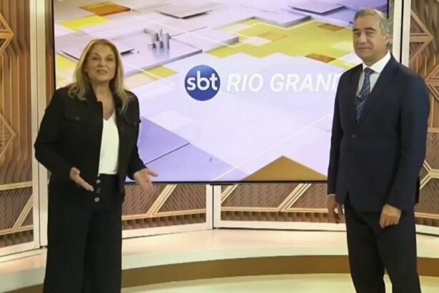 A imagem mostra a dupla de apresentadores Regina Lima e André Haar, em pé, em um estúdio de televisão. Regina, à esquerda, veste um blazer preto, calça preta e uma blusa branca. Ela gesticula com as mãos enquanto fala. André, à direita, usa um terno azul-marinho com gravata. Entre eles, há um grande painel digital exibindo o logotipo "SBT RIO GRANDE".