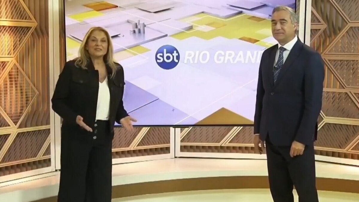 A imagem mostra a dupla de apresentadores Regina Lima e André Haar, em pé, em um estúdio de televisão. Regina, à esquerda, veste um blazer preto, calça preta e uma blusa branca. Ela gesticula com as mãos enquanto fala. André, à direita, usa um terno azul-marinho com gravata. Entre eles, há um grande painel digital exibindo o logotipo "SBT RIO GRANDE".