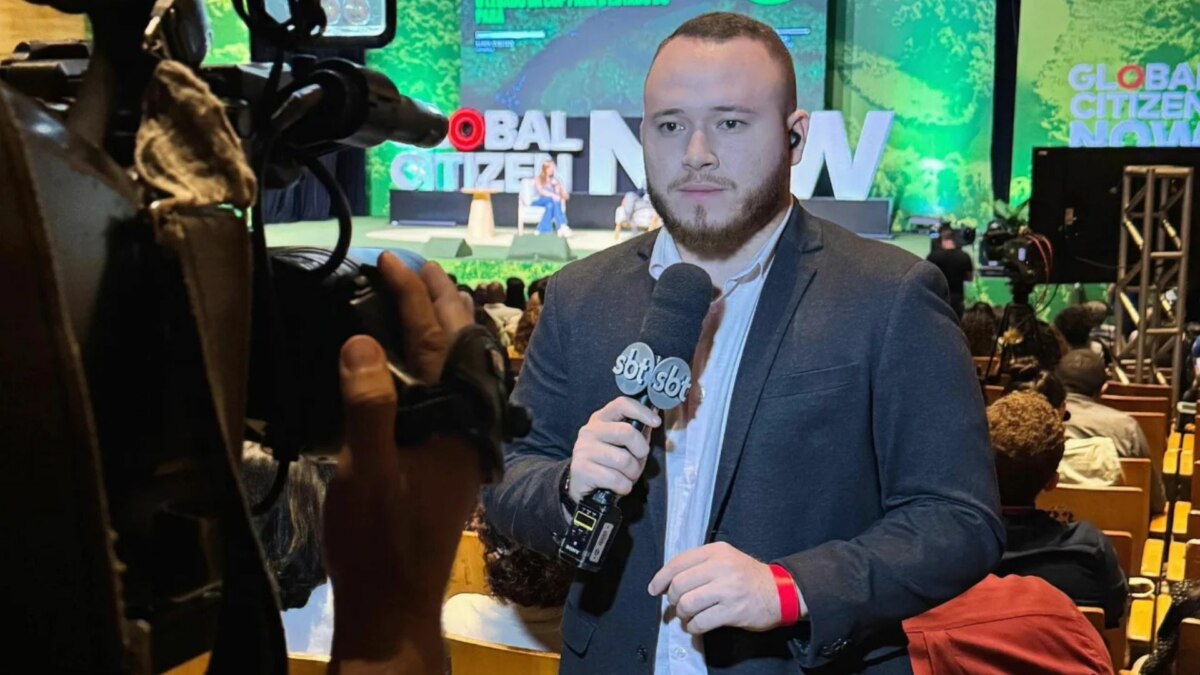 O repórter Andrey Araújo, de terno cinza, segura um microfone com o logo do SBT, enquanto olha para a câmera. Ele está em um evento com um banner 'Global Citizen' no fundo, e o nome do arquivo o associa ao SBT Pará.