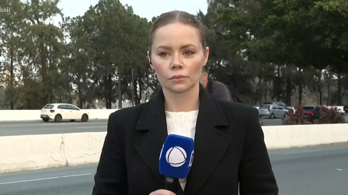 A repórter Beatriz Casadei, de casaco preto, está no acostamento de uma estrada. Ela segura um microfone com a logo da RECORD, e uma expressão séria.