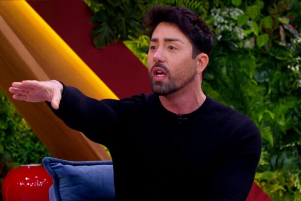 Rico Melquiades, usando um suéter preto, sentado em um sofá no programa "Casos de Família". Ele gesticula com o braço estendido e tem a boca aberta, como se estivesse falando de forma intensa. O fundo é um cenário de estúdio decorado com folhagens e painéis coloridos.