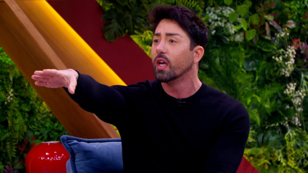 Rico Melquiades, usando um suéter preto, sentado em um sofá no programa "Casos de Família". Ele gesticula com o braço estendido e tem a boca aberta, como se estivesse falando de forma intensa. O fundo é um cenário de estúdio decorado com folhagens e painéis coloridos.