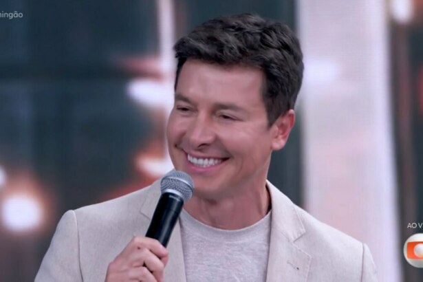 Uma imagem de close-up de Rodrigo Faro, sorrindo e segurando um microfone. Ele usa um blazer claro sobre uma camiseta de tom neutro. Ao fundo, um cenário de estúdio de televisão desfocado. No canto superior esquerdo, a hashtag "#Domingão" e no canto inferior direito, o logo "AO VIVO" da TV Globo.