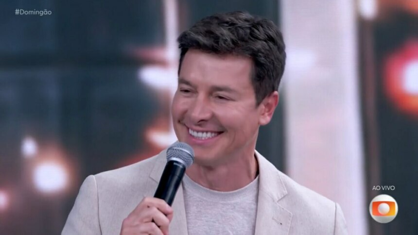 Uma imagem de close-up de Rodrigo Faro, sorrindo e segurando um microfone. Ele usa um blazer claro sobre uma camiseta de tom neutro. Ao fundo, um cenário de estúdio de televisão desfocado. No canto superior esquerdo, a hashtag "#Domingão" e no canto inferior direito, o logo "AO VIVO" da TV Globo.
