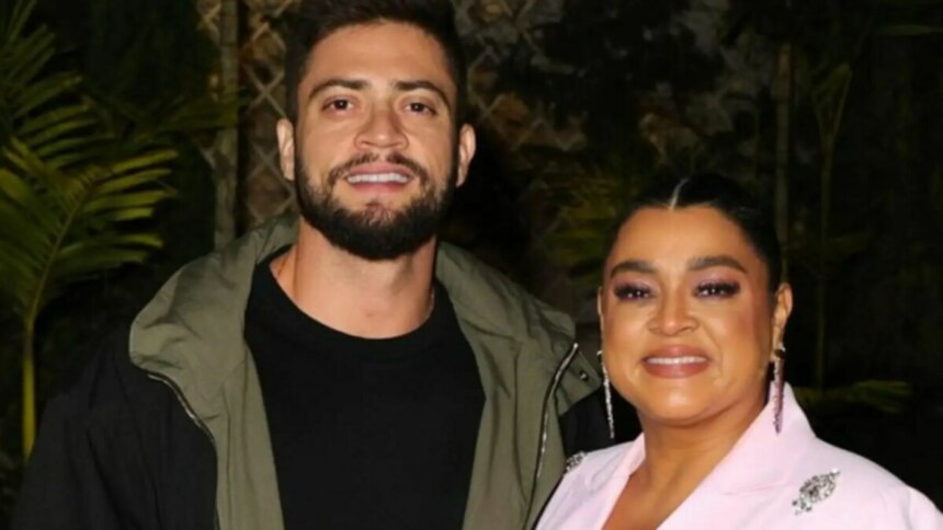 Rodrigo Godoy e Preta Gil posam sorridentes para a foto; ele veste jaqueta verde sobre camiseta preta, e ela usa blazer rosa claro com maquiagem em tons de rosa e brincos longos, em ambiente externo com plantas ao fundo.