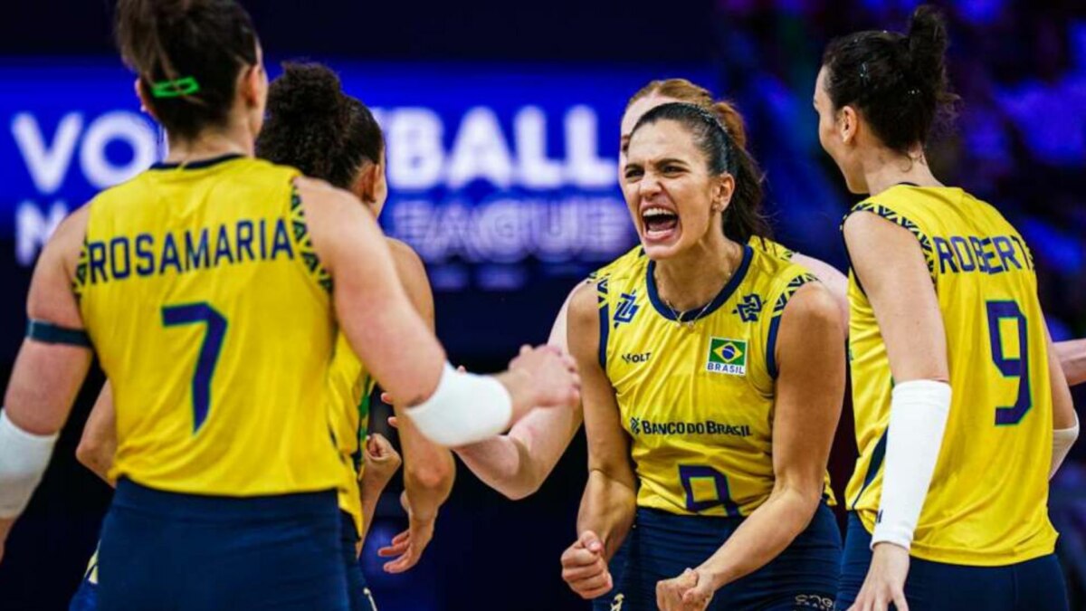 A imagem mostra a jogadora de vôlei Gabi Guimarães, com uma expressão de comemoração, em meio às suas colegas de equipe da Seleção Brasileira. Gabi está vestindo a camisa amarela com detalhes azuis. Ao fundo, uma placa com a inscrição "VOLLEYBALL LEAGUE". As jogadoras Rosamaria e Roberta, com as camisas 7 e 9, respectivamente, também são visíveis na imagem.