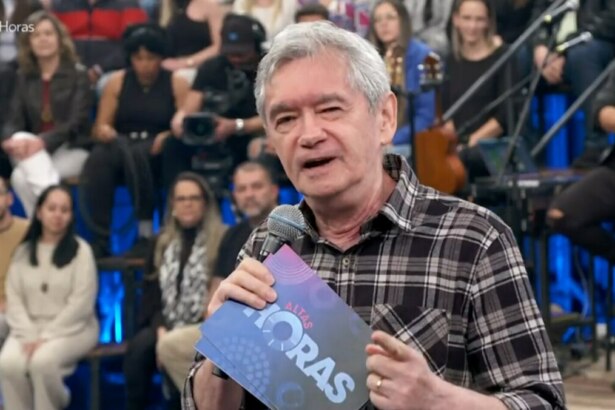 A imagem é um close do apresentador Serginho Groisman, no palco do programa "Altas Horas". Ele está sorrindo, olhando para o lado, e segura um microfone com a logo do programa e um pequeno tablet. Ele veste uma camisa xadrez escura. A plateia está ao fundo, sentada em uma arquibancada com luzes azuis.