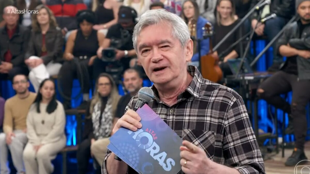 A imagem é um close do apresentador Serginho Groisman, no palco do programa "Altas Horas". Ele está sorrindo, olhando para o lado, e segura um microfone com a logo do programa e um pequeno tablet. Ele veste uma camisa xadrez escura. A plateia está ao fundo, sentada em uma arquibancada com luzes azuis.