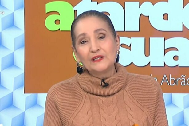 A apresentadora Sonia Abrão em um estúdio de televisão, falando diretamente para a câmera. Ela usa um suéter de gola alta na cor caramelo e grandes brincos. Ao fundo, o painel do cenário exibe o nome do programa "A Tarde é Sua" e o nome dela.
