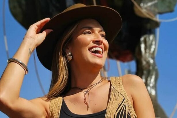 A imagem é um close da apresentadora Talitha Morete durante a Festa do Peão de Barretos. Ela está sorrindo e olhando para o lado. Ela usa um chapéu de caubói marrom, uma blusa preta, um colete de camurça com franjas e brincos prateados. O fundo é um céu azul claro e uma escultura escura e desfocada.