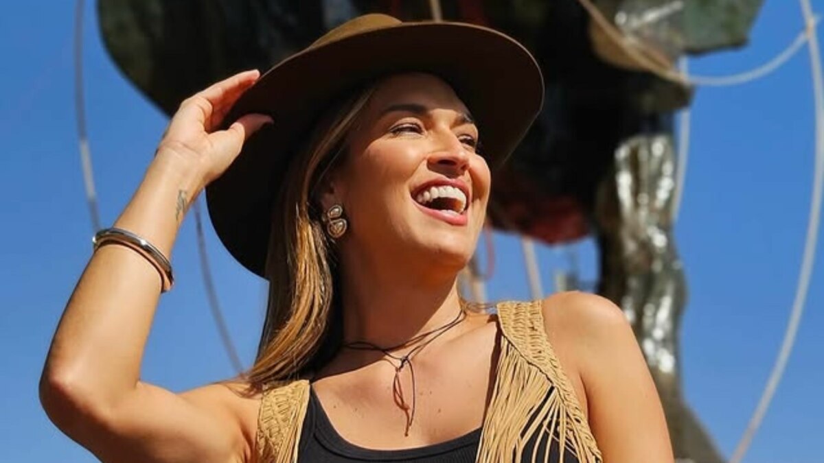 A imagem é um close da apresentadora Talitha Morete durante a Festa do Peão de Barretos. Ela está sorrindo e olhando para o lado. Ela usa um chapéu de caubói marrom, uma blusa preta, um colete de camurça com franjas e brincos prateados. O fundo é um céu azul claro e uma escultura escura e desfocada.