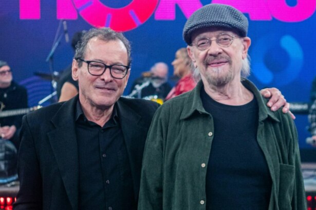 Dois integrantes da banda The Fevers posam no palco do programa "Altas Horas". À esquerda, um homem de óculos e blazer preto sorri para a câmera. Ao lado dele, um homem mais velho com barba, óculos e uma boina, sorri e tem o braço esquerdo sobre o ombro do colega. Ao fundo, parcialmente visíveis, outros músicos e equipamentos de palco em um ambiente com iluminação azulada e elementos do cenário do programa.