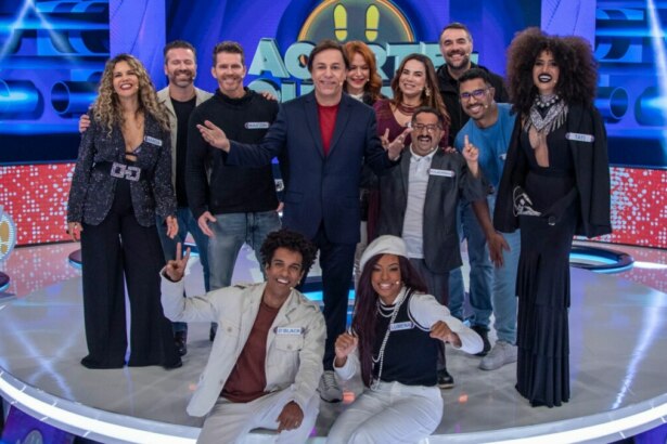 Tom Cavalcante e parte do elenco do programa "Acerte ou Caia" posam juntos em um palco iluminado. Tom, no centro, veste um terno escuro com camisa vermelha, sorrindo e apontando. Ao redor dele, diversas pessoas com figurinos variados, incluindo um homem de blazer branco agachado à frente, e uma mulher com cabelo encaracolado e roupa preta. O cenário futurista é azul com luzes vibrantes e detalhes circulares, e o público pode ser visto ao fundo.