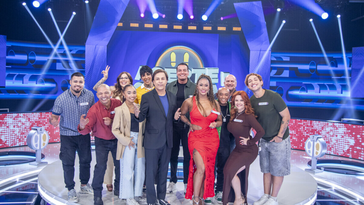 apresentador Tom Cavalcante e o elenco do programa 'Acerte ou Caia' posam para uma foto de grupo em um palco de TV. Tom está no centro, de blazer preto, ao lado de uma mulher em um vestido vermelho com uma fenda alta.
