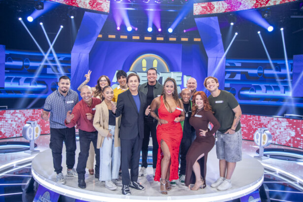 apresentador Tom Cavalcante e o elenco do programa 'Acerte ou Caia' posam para uma foto de grupo em um palco de TV. Tom está no centro, de blazer preto, ao lado de uma mulher em um vestido vermelho com uma fenda alta.