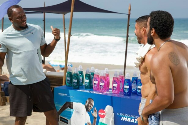 A imagem mostra Usain Bolt em uma praia. Ele está de costas para a câmera, gesticulando com as mãos enquanto conversa com dois homens, um dos quais é o ator Cauã Reymond. Usain Bolt veste uma camiseta clara e shorts escuros. No centro da imagem, há um estande com garrafas de produtos OMO em várias cores e uma imagem de Usain Bolt na frente. O ator Cauã Reymond, à direita, está sem camisa e com uma toalha branca no ombro. Ao fundo, é possível ver o mar e a areia da praia, com algumas cadeiras de praia e guarda-sóis. A cena é da novela Vale Tudo na Globo.