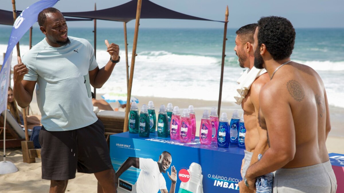 A imagem mostra Usain Bolt em uma praia. Ele está de costas para a câmera, gesticulando com as mãos enquanto conversa com dois homens, um dos quais é o ator Cauã Reymond. Usain Bolt veste uma camiseta clara e shorts escuros. No centro da imagem, há um estande com garrafas de produtos OMO em várias cores e uma imagem de Usain Bolt na frente. O ator Cauã Reymond, à direita, está sem camisa e com uma toalha branca no ombro. Ao fundo, é possível ver o mar e a areia da praia, com algumas cadeiras de praia e guarda-sóis. A cena é da novela Vale Tudo na Globo.