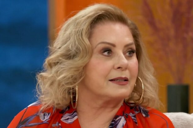 A imagem é um close da atriz Vera Fischer em uma entrevista ao Encontro. Ela está sentada, usando uma blusa vermelha com estampa floral e grandes brincos de argola dourados. O cabelo loiro dela está arrumado em cachos e ela tem uma expressão séria. O fundo da imagem está desfocado, com tons de laranja e azul.