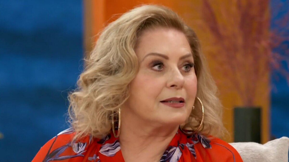 A imagem é um close da atriz Vera Fischer em uma entrevista ao Encontro. Ela está sentada, usando uma blusa vermelha com estampa floral e grandes brincos de argola dourados. O cabelo loiro dela está arrumado em cachos e ela tem uma expressão séria. O fundo da imagem está desfocado, com tons de laranja e azul.