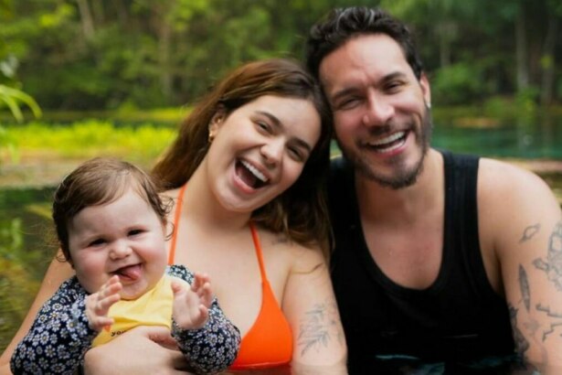A influenciadora Viih Tube e seu marido, Eliezer, seguram a filha Lua em um rio ou lagoa. Os três sorriem para a câmera. Viih usa um top de biquíni laranja, Eliezer uma regata preta, e a bebê Lua usa uma blusa amarela com estampa floral. Ao fundo, uma mata densa.