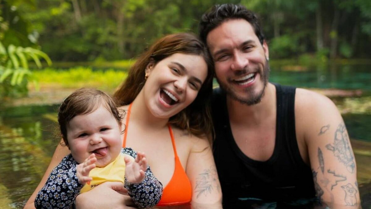 A influenciadora Viih Tube e seu marido, Eliezer, seguram a filha Lua em um rio ou lagoa. Os três sorriem para a câmera. Viih usa um top de biquíni laranja, Eliezer uma regata preta, e a bebê Lua usa uma blusa amarela com estampa floral. Ao fundo, uma mata densa.