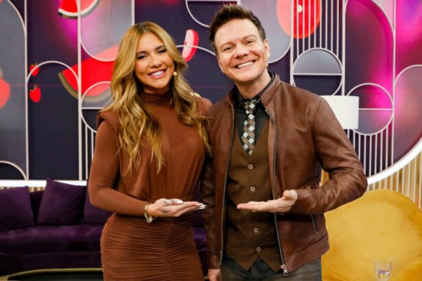 Virginia Fonseca e Michel Teló posam sorridentes no cenário do programa "Sabadou". Virginia veste um vestido marrom justo, enquanto Michel Teló usa uma camisa, colete e jaqueta de couro marrons, com uma gravata estampada. Ambos estendem as mãos abertas para a frente. O cenário possui tons de roxo, rosa e dourado, com formas geométricas e sofás roxos e amarelos.