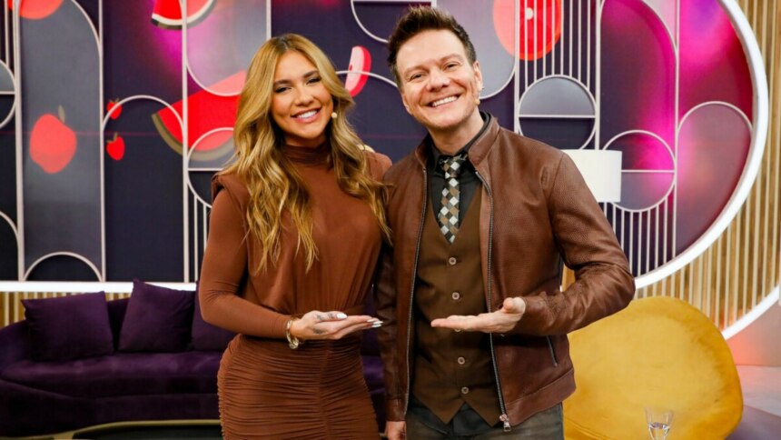 Virginia Fonseca e Michel Teló posam sorridentes no cenário do programa "Sabadou". Virginia veste um vestido marrom justo, enquanto Michel Teló usa uma camisa, colete e jaqueta de couro marrons, com uma gravata estampada. Ambos estendem as mãos abertas para a frente. O cenário possui tons de roxo, rosa e dourado, com formas geométricas e sofás roxos e amarelos.