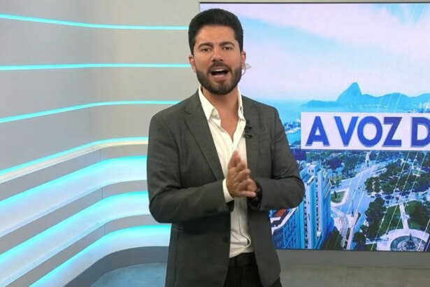 O apresentador Wagner Montes Filho em um estúdio de TV. Ele usa um paletó cinza e uma camisa branca com a gola aberta. Ele gesticula com as mãos e olha para a câmera. Ao fundo, há um painel digital com a vista de uma cidade.