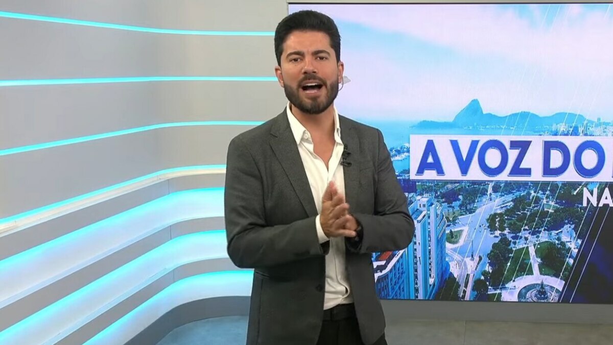O apresentador Wagner Montes Filho em um estúdio de TV. Ele usa um paletó cinza e uma camisa branca com a gola aberta. Ele gesticula com as mãos e olha para a câmera. Ao fundo, há um painel digital com a vista de uma cidade.