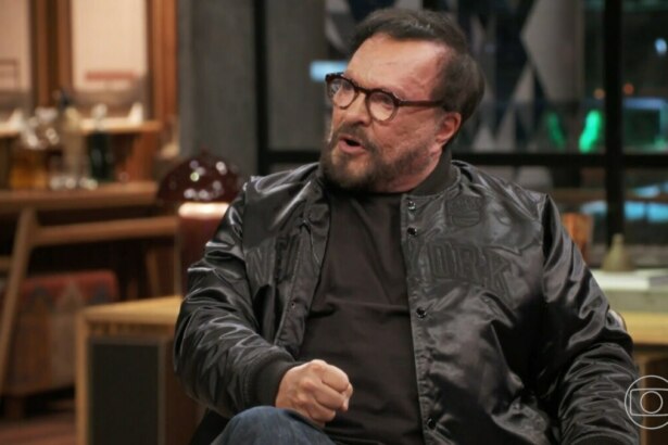 A imagem é um close do diretor e ator Wolf Maya, em uma entrevista no programa "Conversa com Bial". Ele está sentado, com a boca aberta como se estivesse falando, e olha para o lado. Ele usa óculos de armação escura, uma jaqueta preta de material brilhante sobre uma camiseta preta. A barba e o cabelo dele são escuros. O fundo da cena está desfocado e mostra um ambiente interno, com o logo da Globo no canto inferior direito.