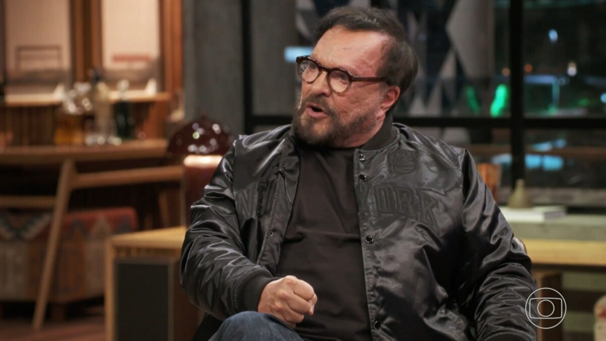 A imagem é um close do diretor e ator Wolf Maya, em uma entrevista no programa "Conversa com Bial". Ele está sentado, com a boca aberta como se estivesse falando, e olha para o lado. Ele usa óculos de armação escura, uma jaqueta preta de material brilhante sobre uma camiseta preta. A barba e o cabelo dele são escuros. O fundo da cena está desfocado e mostra um ambiente interno, com o logo da Globo no canto inferior direito.