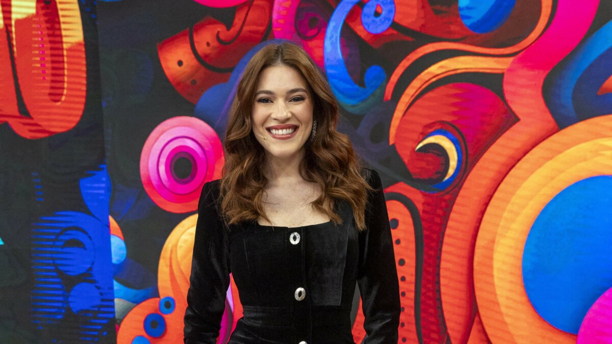 A apresentadora Ana Clara sorri em frente a um fundo vibrante e colorido no cenário da Dança dos Famosos do Domingão. Ela veste um casaco de veludo preto.