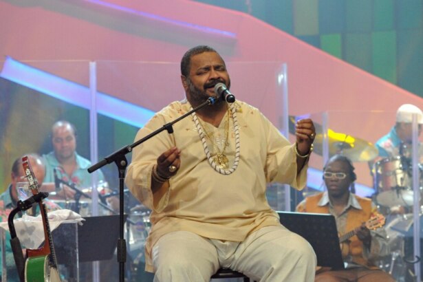 Arlindo Cruz canta sentado em um palco, vestindo roupa clara e colares, enquanto segura o microfone. Ao fundo, músicos tocam instrumentos.