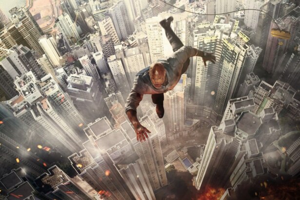 Cena do filme “Arranha-Céu: Coragem Sem Limite” mostrando o personagem de Dwayne Johnson despencando de uma grua no topo de um arranha-céu em chamas, com a cidade ao fundo e fumaça densa subindo do incêndio.