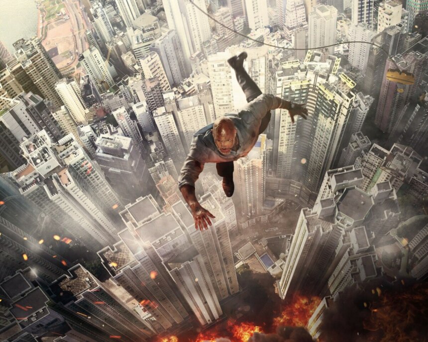 Cena do filme “Arranha-Céu: Coragem Sem Limite” mostrando o personagem de Dwayne Johnson despencando de uma grua no topo de um arranha-céu em chamas, com a cidade ao fundo e fumaça densa subindo do incêndio.