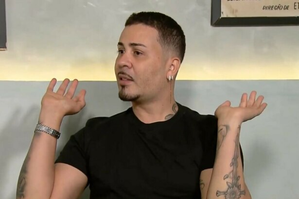 Carlinhos Maia em uma entrevista ao Domingo Legal no SBT, com as mãos levantadas e as palmas viradas para cima, em um gesto de surpresa ou dúvida. Ele usa uma camiseta preta e um relógio prateado no pulso esquerdo. O influenciador tem tatuagens nos braços, barba e cabelo curtos.