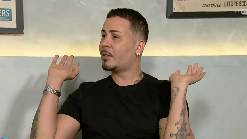 Carlinhos Maia em uma entrevista ao Domingo Legal no SBT, com as mãos levantadas e as palmas viradas para cima, em um gesto de surpresa ou dúvida. Ele usa uma camiseta preta e um relógio prateado no pulso esquerdo. O influenciador tem tatuagens nos braços, barba e cabelo curtos.