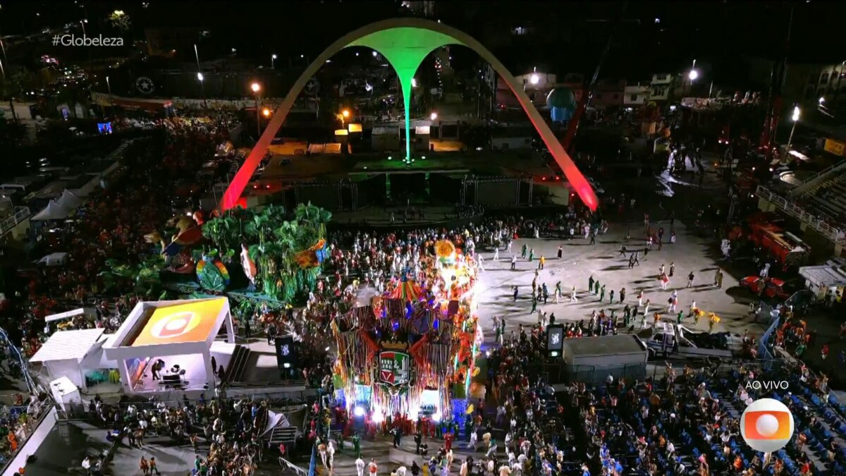 Imagem aérea noturna do Sambódromo do Rio de Janeiro durante um desfile de carnaval. A passarela está cheia de passistas e alegorias de uma escola de samba. As arquibancadas estão repletas de público. O icônico arco central está iluminado em vermelho e verde. A hashtag "#Globeleza" e o logo da Globo com a indicação "AO VIVO" são visíveis