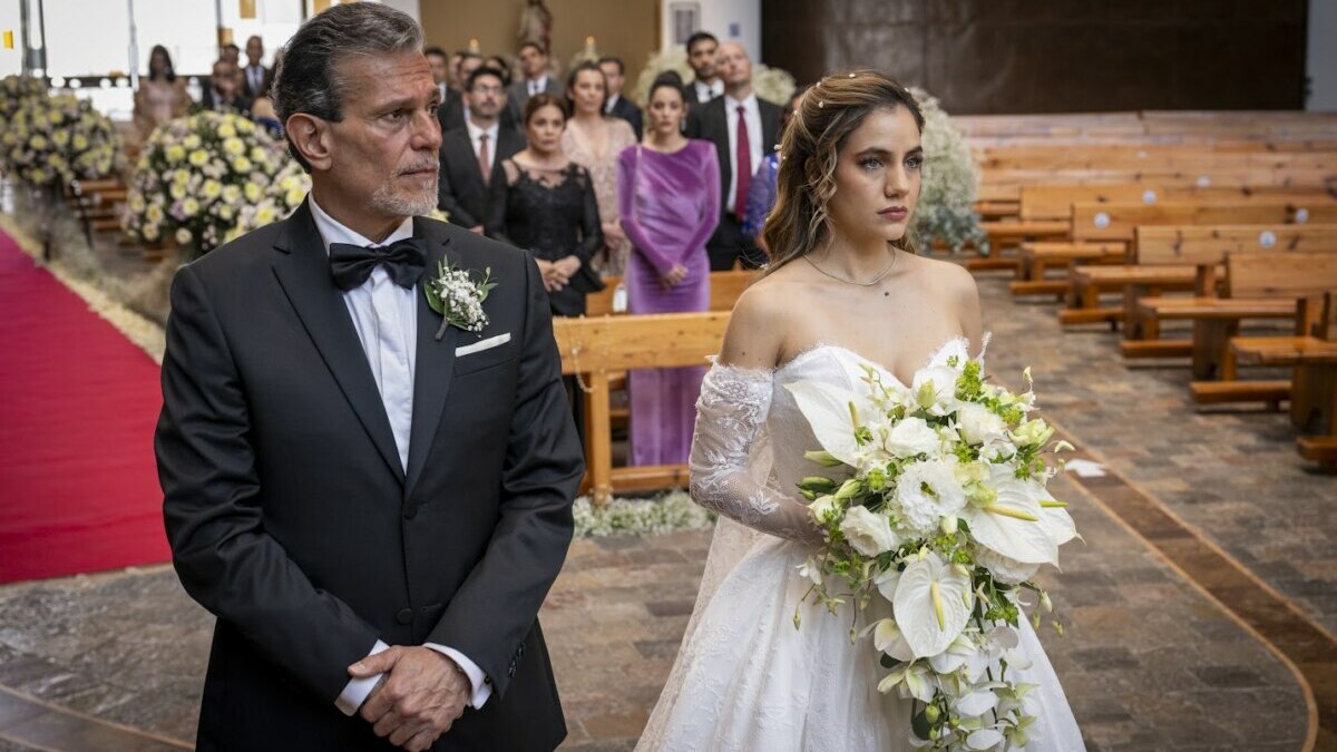 Cena do casamento de Mar em As Filhas da Senhora Garcia (SBT), mostrando a noiva com vestido branco e buquê de flores ao lado de um homem de terno preto no altar, com convidados ao fundo