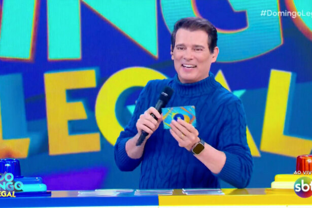 O apresentador Celso Portiolli sorrindo, segurando um cartão azul com uma bolinha amarela, no programa Domingo Legal. Ele veste um suéter azul de gola alta e um relógio de pulso. No fundo, a tela exibe o logo "Domingo Legal" e as hashtags "#DomingoLegal" e "#SBT", com a indicação "AO VIVO". No canto inferior esquerdo, uma bancada azul e amarela é visível.