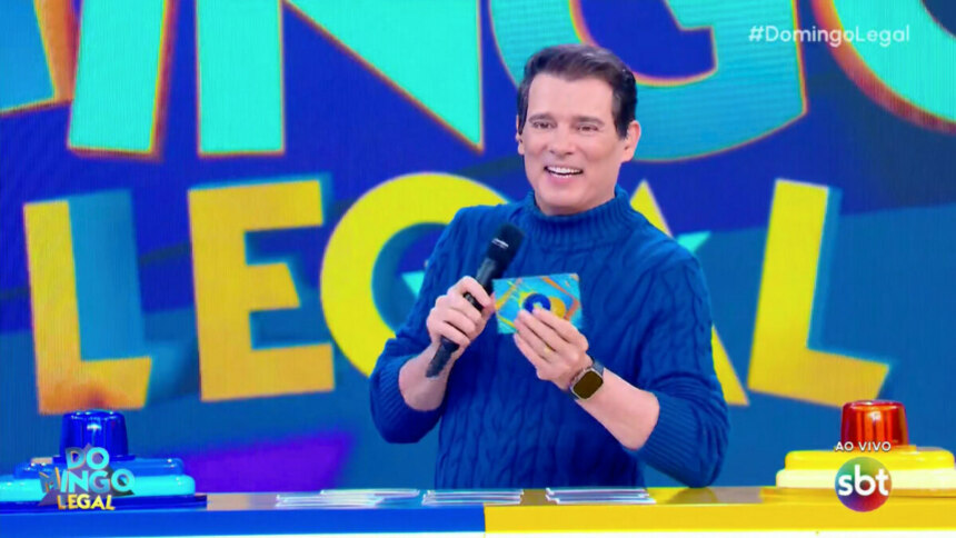 O apresentador Celso Portiolli sorrindo, segurando um cartão azul com uma bolinha amarela, no programa Domingo Legal. Ele veste um suéter azul de gola alta e um relógio de pulso. No fundo, a tela exibe o logo "Domingo Legal" e as hashtags "#DomingoLegal" e "#SBT", com a indicação "AO VIVO". No canto inferior esquerdo, uma bancada azul e amarela é visível.