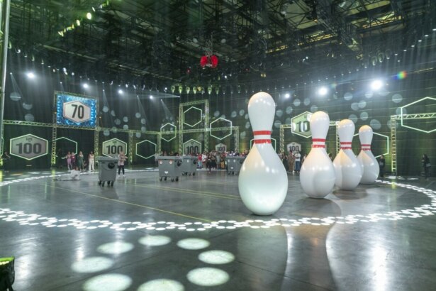 Cenário do programa Game dos 100 com 70 competidores ao fundo, grandes pinos de boliche infláveis posicionados no centro da arena e caçambas que serão usadas na prova do boliche gigante, com iluminação e painéis com o número 100 ao redor do estúdio.