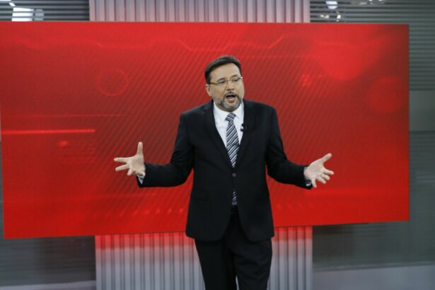 Geraldo Luís na estreia do "Aqui Agora" em 2025, no palco com um fundo de LED vermelho, usando um terno preto, camisa branca e gravata listrada, gesticulando com as mãos abertas enquanto se dirige ao público
