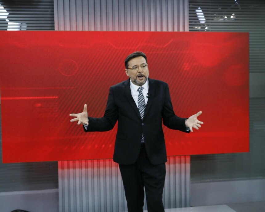 Geraldo Luís na estreia do "Aqui Agora" em 2025, no palco com um fundo de LED vermelho, usando um terno preto, camisa branca e gravata listrada, gesticulando com as mãos abertas enquanto se dirige ao público